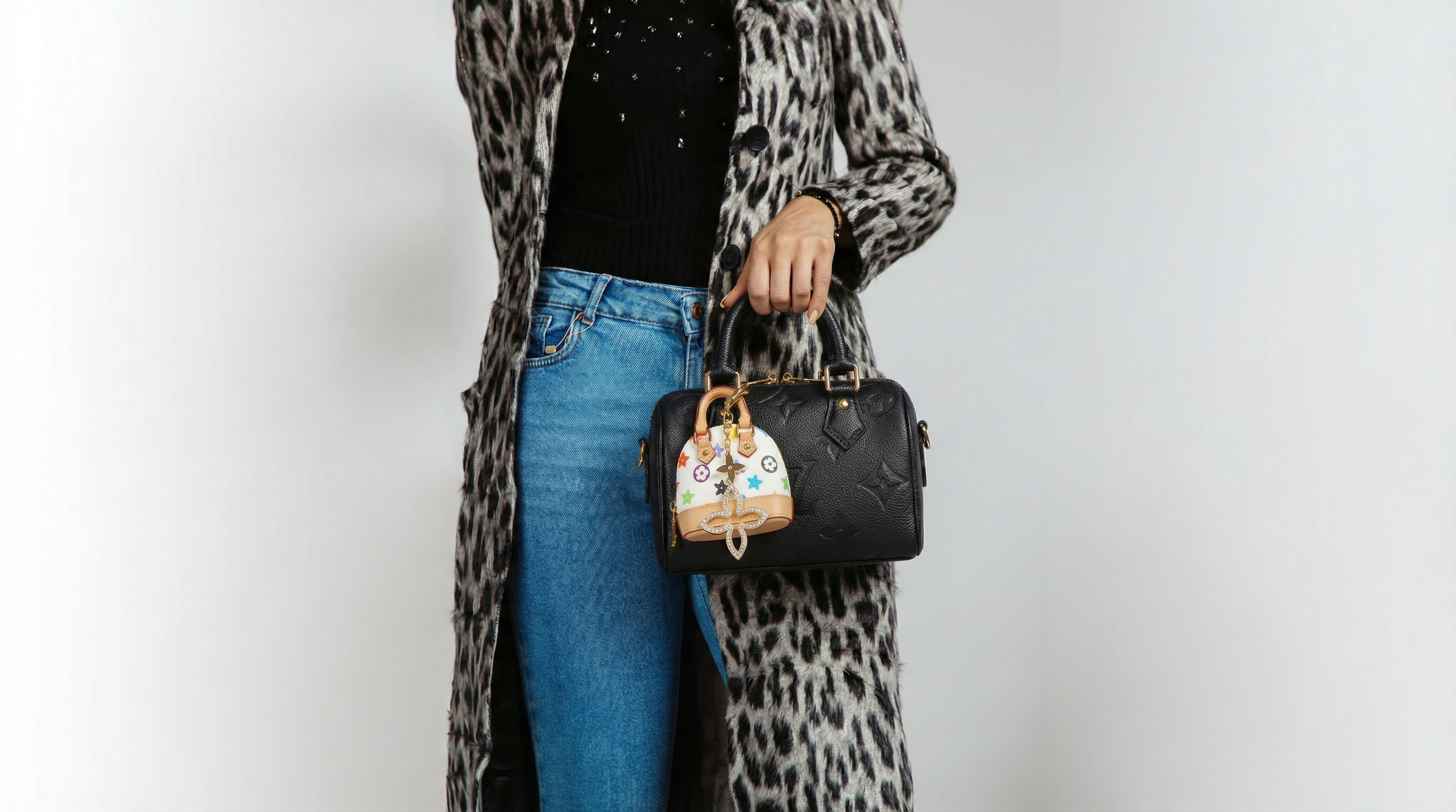Person wearing a leopard print coat, blue jeans, and holding a black handbag with a colorful icon handbag charm on a white background